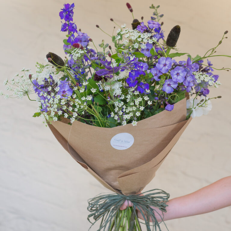 A hand holds the Hand-tied Bouquet of Seasonal Flowers, featuring purple, white, and green blooms wrapped in brown paper with a rustic string tie.