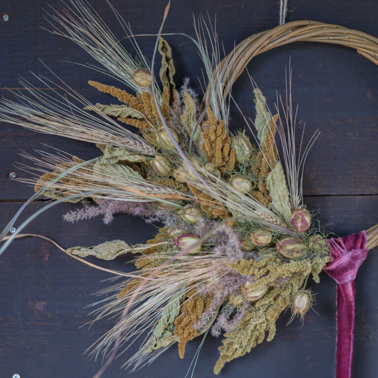 The Twelve Drummers Drumming wreath, fashioned from dried grasses, foliage, and seed pods, hangs against a dark wooden background with a purple ribbon.