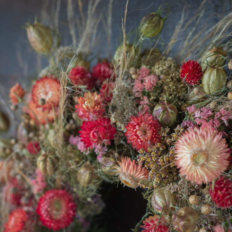 The "Five Golllldddddddddd Rings" is a wreath crafted from a variety of dried flowers and grasses, showcasing shades of red, pink, and orange for a textured, rustic aesthetic.