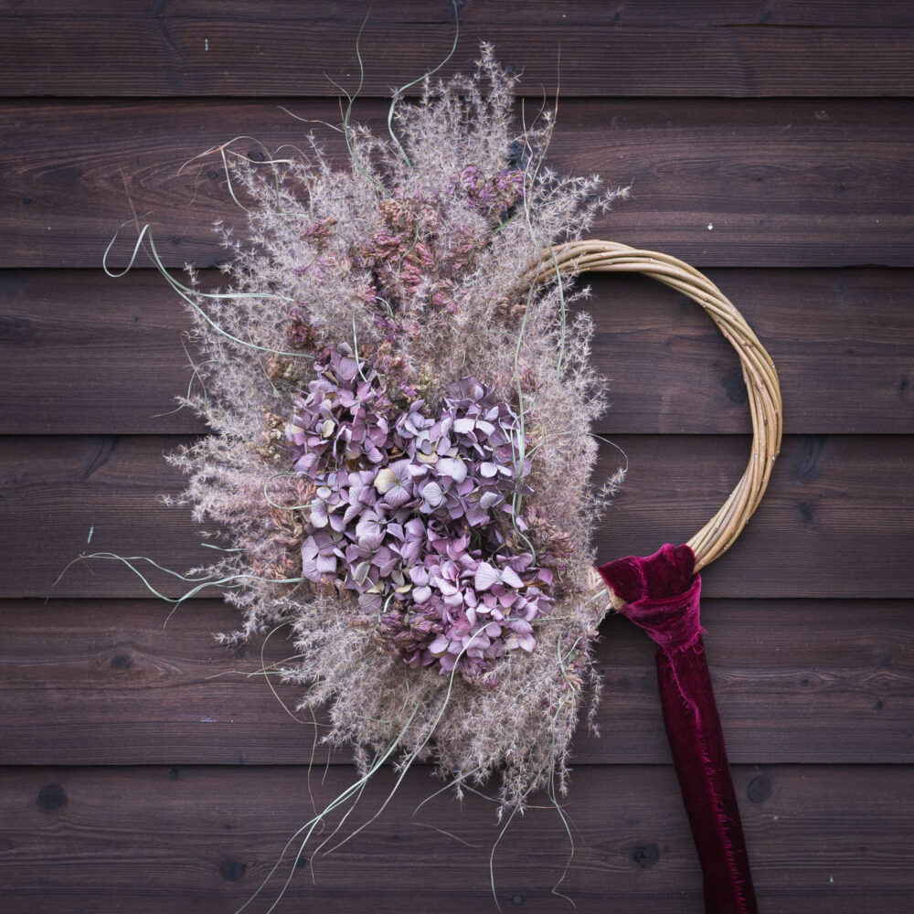 The Twelve Drummers Drumming wreath, featuring dried hydrangeas and grasses with a red velvet ribbon, is beautifully displayed on a wooden wall.
