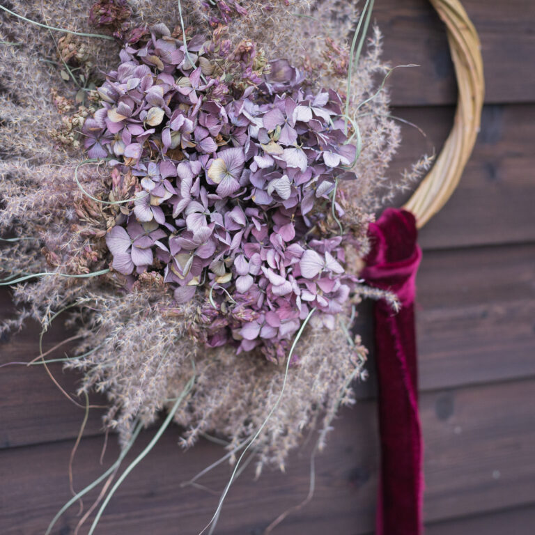 The "Wreath - Twelve Drummers Drumming" showcases a circle of purple hydrangeas and dried grasses, finished with a maroon ribbon, all set against a wooden background.