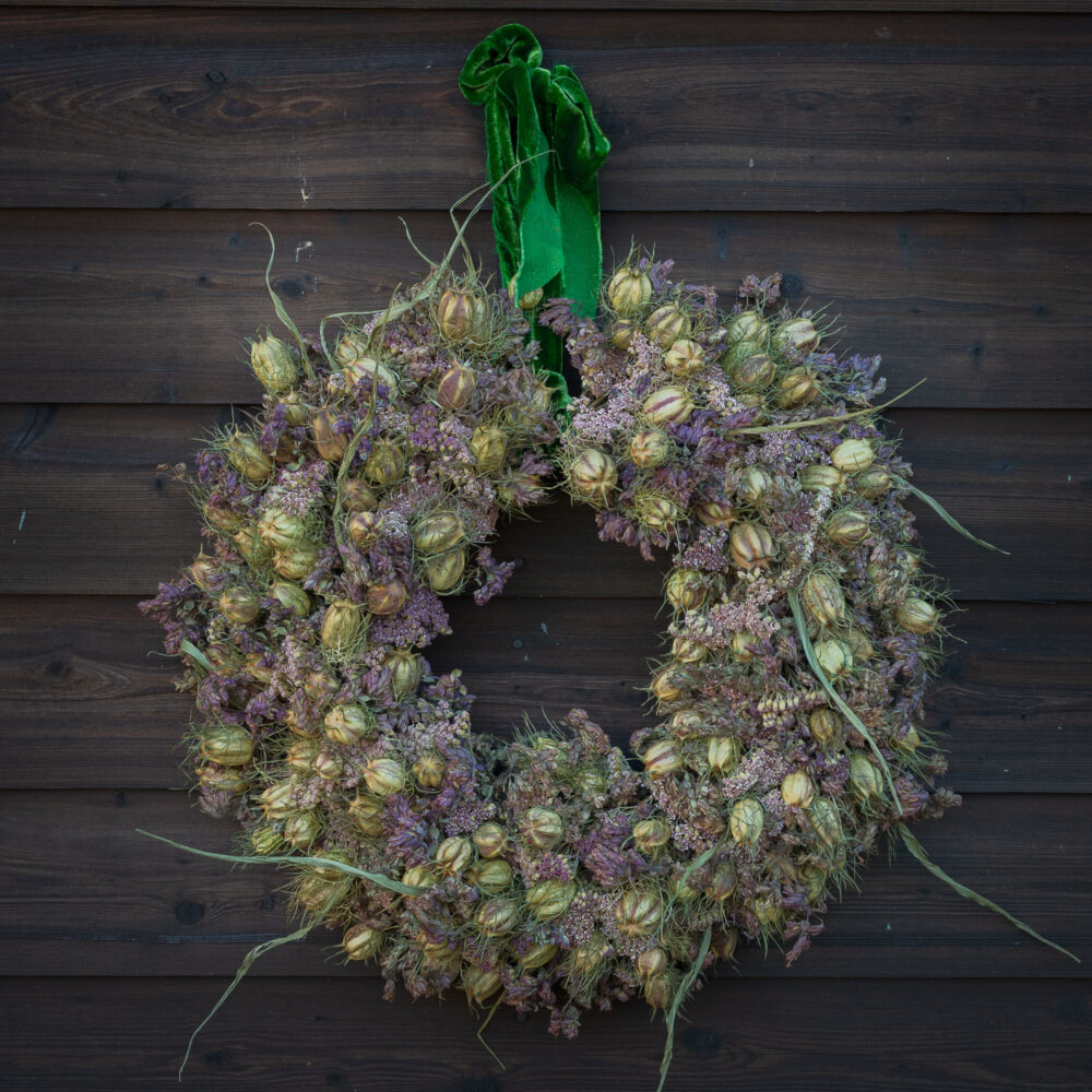 The "Twelve Drummers Drumming" wreath, crafted from dried flowers and seed pods with a green ribbon, adorns a dark wooden wall.