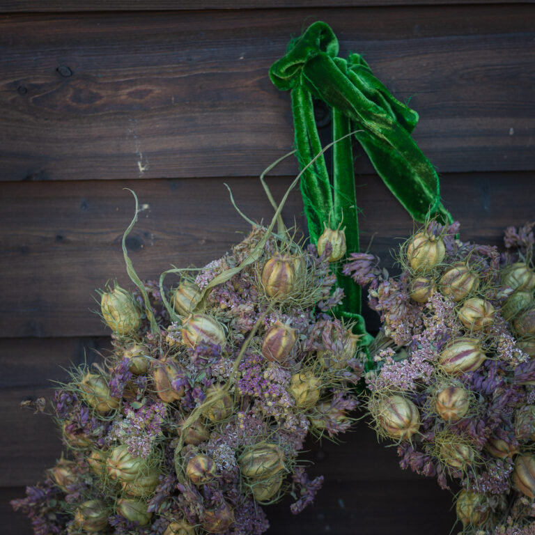 The "Wreath - Twelve Drummers Drumming," showcasing purple and beige dried flowers, is elegantly displayed on a dark wooden wall with a green velvet ribbon.