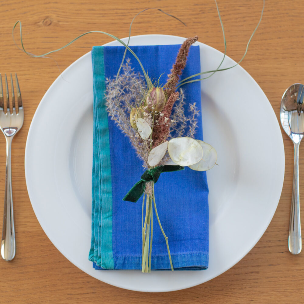 A Table Place-Setting - Blue & Purple comprises a white plate with a blue serviette and decorative dried flowers, featuring a silver fork on the left and a spoon on the right.