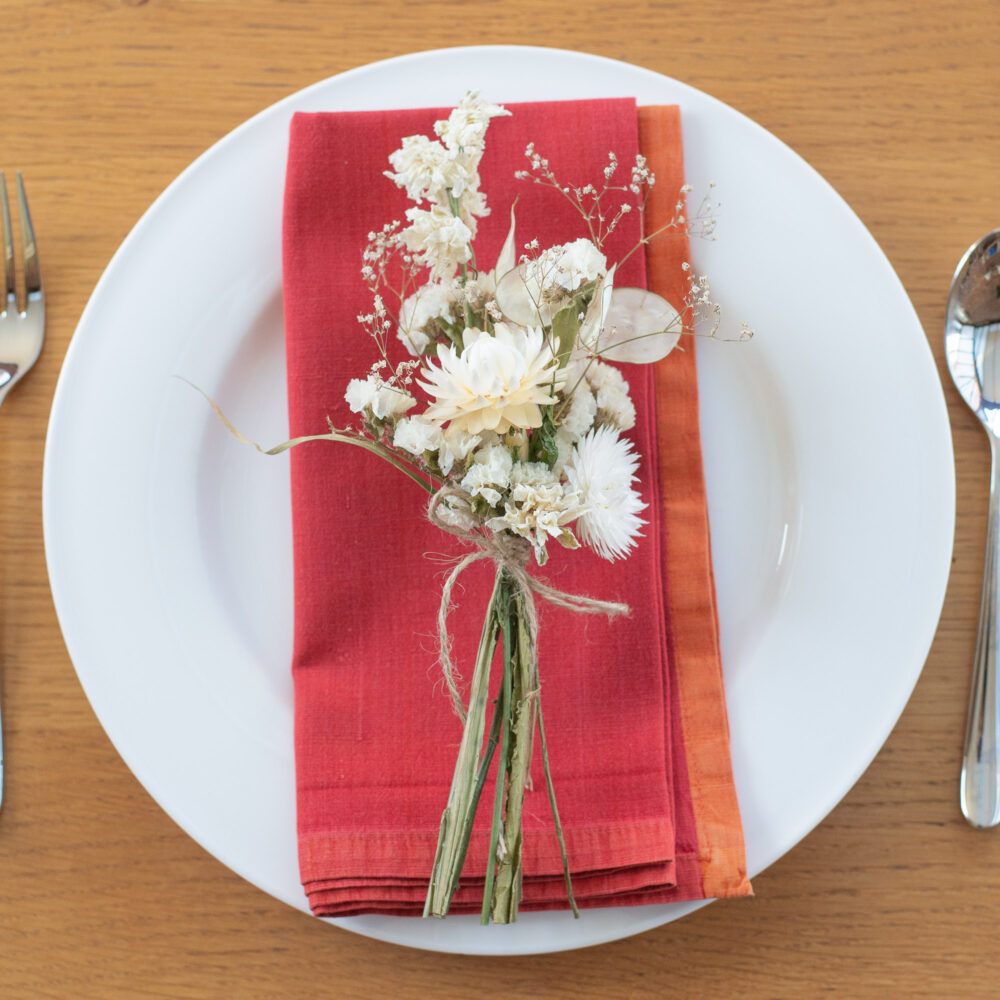 A blue and purple table place-setting features a neatly arranged plate on a wooden table, accented by folded serviettes. A small bouquet of white flowers tied with twine decorates the setting, with cutlery placed on either side.