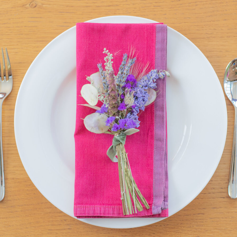 A Table Place-Setting - Blue and Purple on a wooden table, featuring a pink serviette with purple flowers on a white plate, and cutlery arranged alongside.