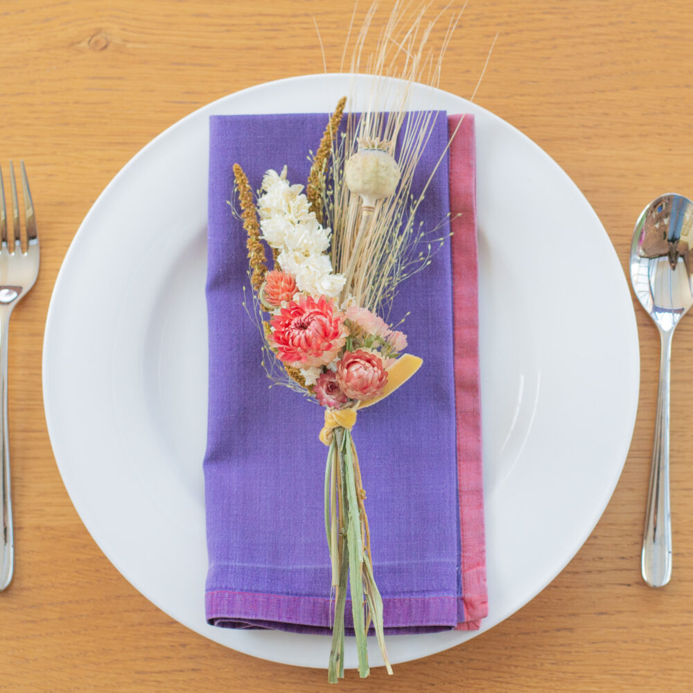 A white plate rests on a wooden table adorned with a purple serviette and a small dried floral bouquet, including wheat and pink flowers. A fork and spoon are placed beside it as part of the Table Place-Setting - Blue & Purple.