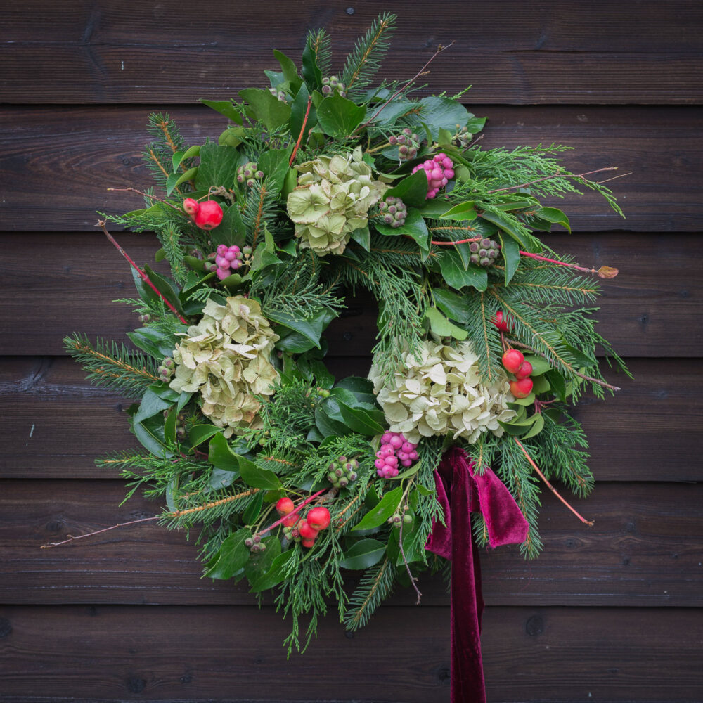 The Twelve Drummers Drumming wreath features greenery, hydrangeas, red berries, and a red ribbon on a wooden wall.