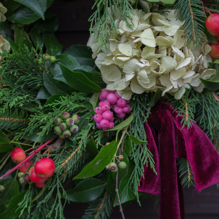 The "Eight Maids-a-Milking" wreath features green leaves, red and pink berries, off-white dried hydrangeas, evergreen sprigs, and a burgundy velvet ribbon.