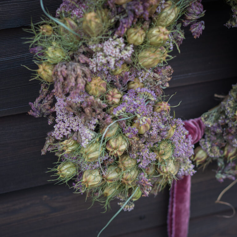The "Twelve Drummers Drumming" wreath, crafted from dried flowers and seed pods in purple and green hues, adorns a dark wooden wall with a velvet ribbon hanging at the bottom.