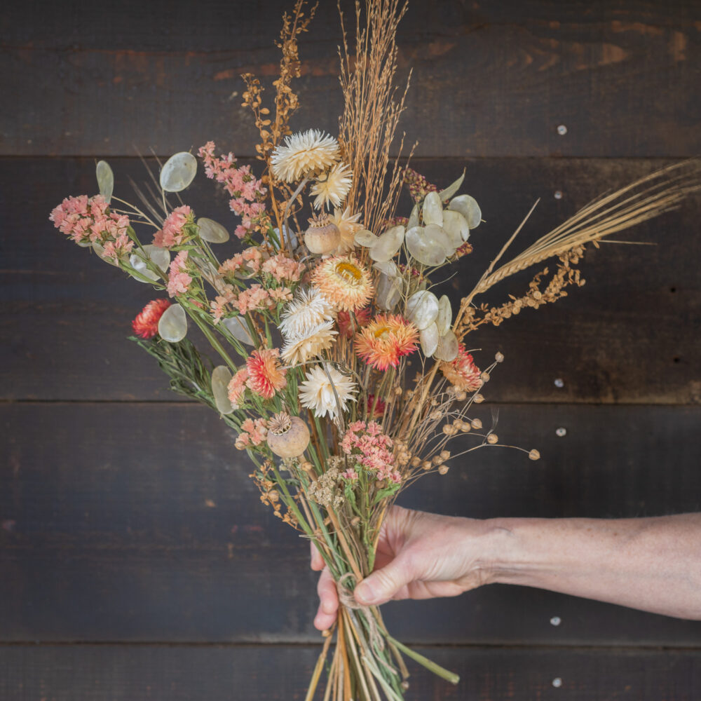 A hand holding the "Dried Flower Bouquet - Bright" against a dark wooden backdrop.