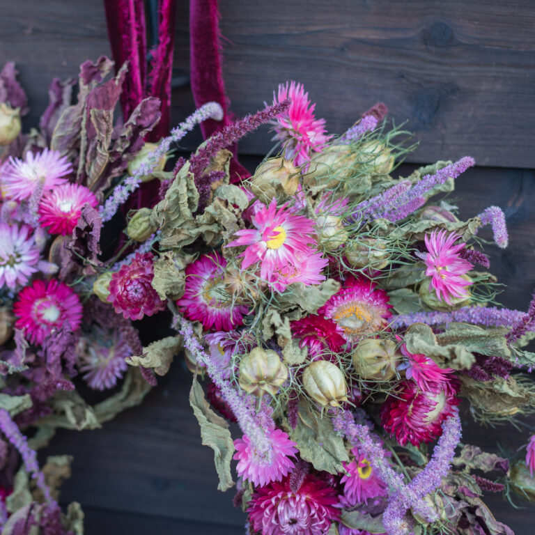 The "Dried Flower Bouquet - Bright," featuring mainly pink and purple dried flowers with a variety of textures and shapes, hangs against a dark wooden background.