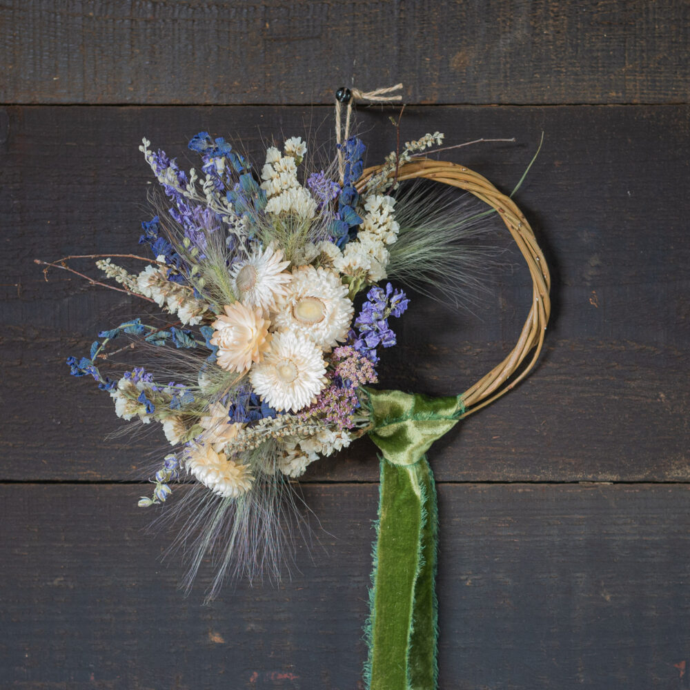 The Mini Wreath - Green & White features a round wicker base decorated with white and purple dried flowers, green foliage, and a green velvet ribbon, set against a dark wooden background.