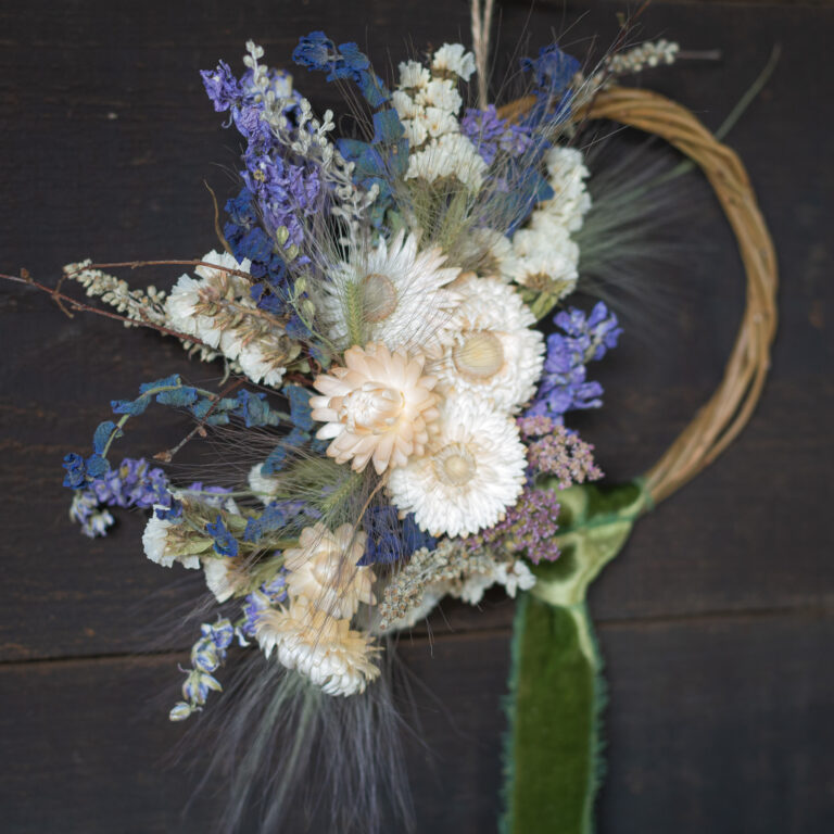The "Mini Wreath - Green & White" showcases a dried floral arrangement of white and purple flowers, green foliage, and a twisted brown base, set against a dark wooden background.