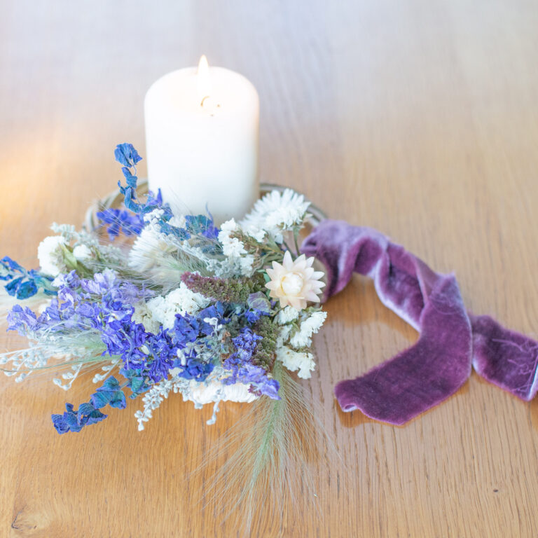 A lit white candle sits beside a green and white mini wreath adorned with blue and white flowers, tied with a purple velvet ribbon, on a wooden surface.
