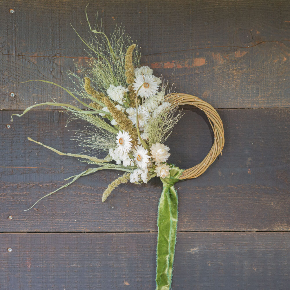 The Mini Wreath - Green & White, featuring white flowers and green foliage with a green ribbon, adorns a dark wooden wall.