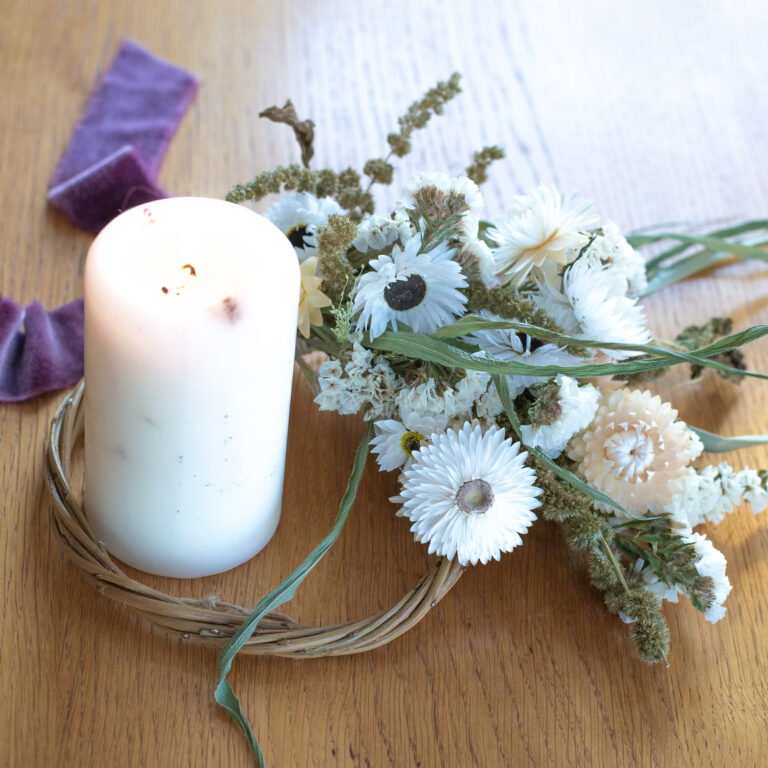 A Mini Wreath - Green & White, featuring white flowers, is arranged on a wooden surface with a white pillar candle and a nearby purple ribbon.