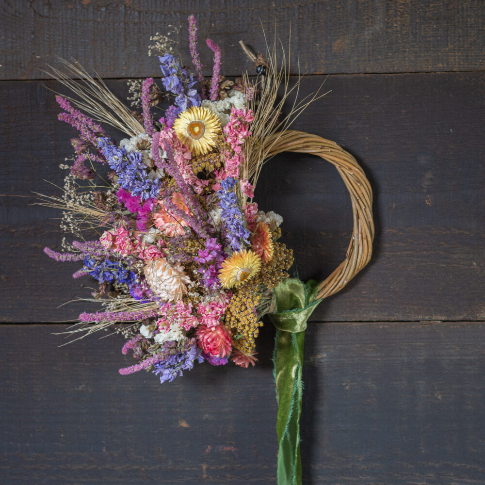 A Mini Wreath - Green & White, featuring a curved handle adorned with colourful dried flowers and grasses, is set against a dark wooden background.