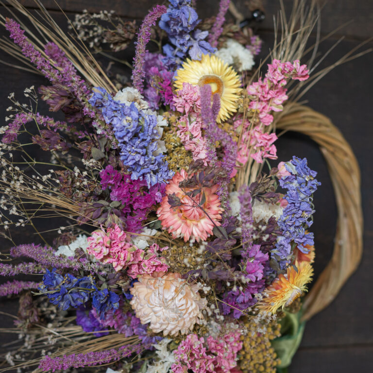 A vibrant Mini Wreath - Green & White, featuring a display of dried flowers in pink, purple, yellow, and white arranged in a spiral against a dark background.