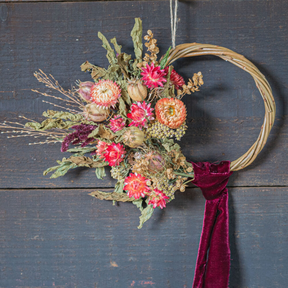 A Mini Wreath - Green & White, crafted from dried flowers and foliage with a dark red velvet ribbon, hangs against a weathered wooden wall.