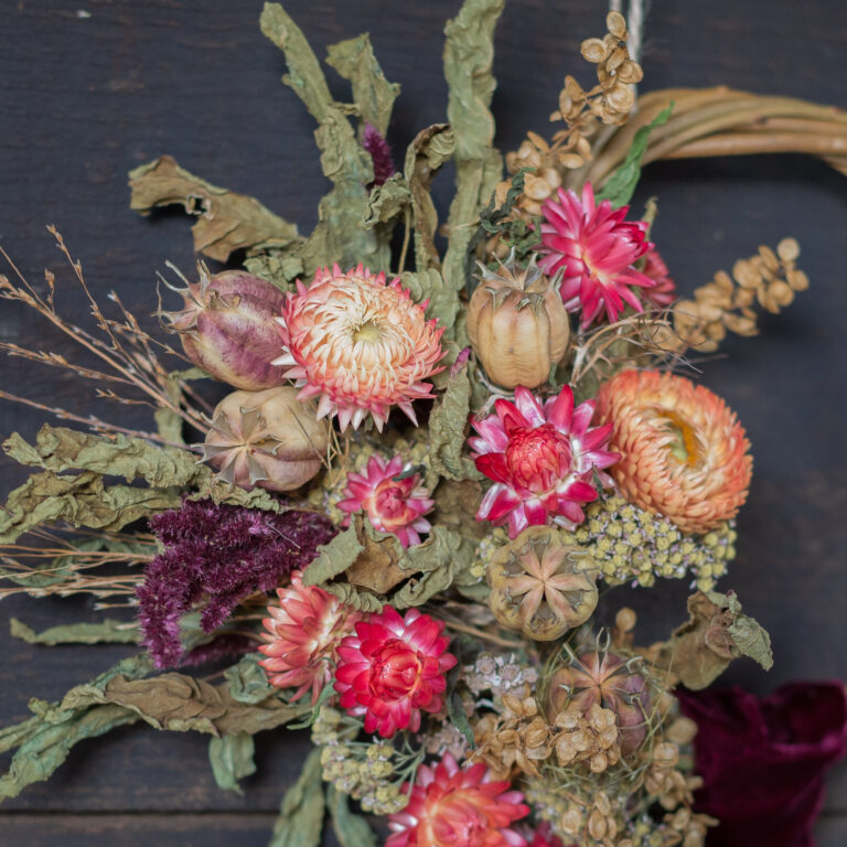 A Mini Wreath - Green & White featuring pink and orange dried flowers, brown seed pods, and green leaves set against a dark background.