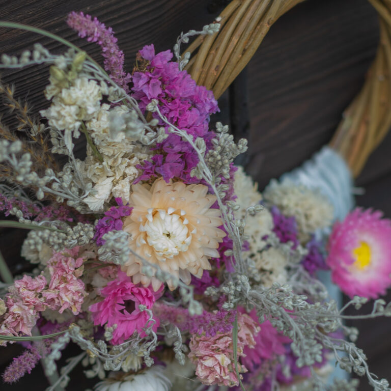 A close-up of the Mini Wreath - Pastels displays pink, purple, cream, and white dried flowers with assorted leaves against a dark wooden background.