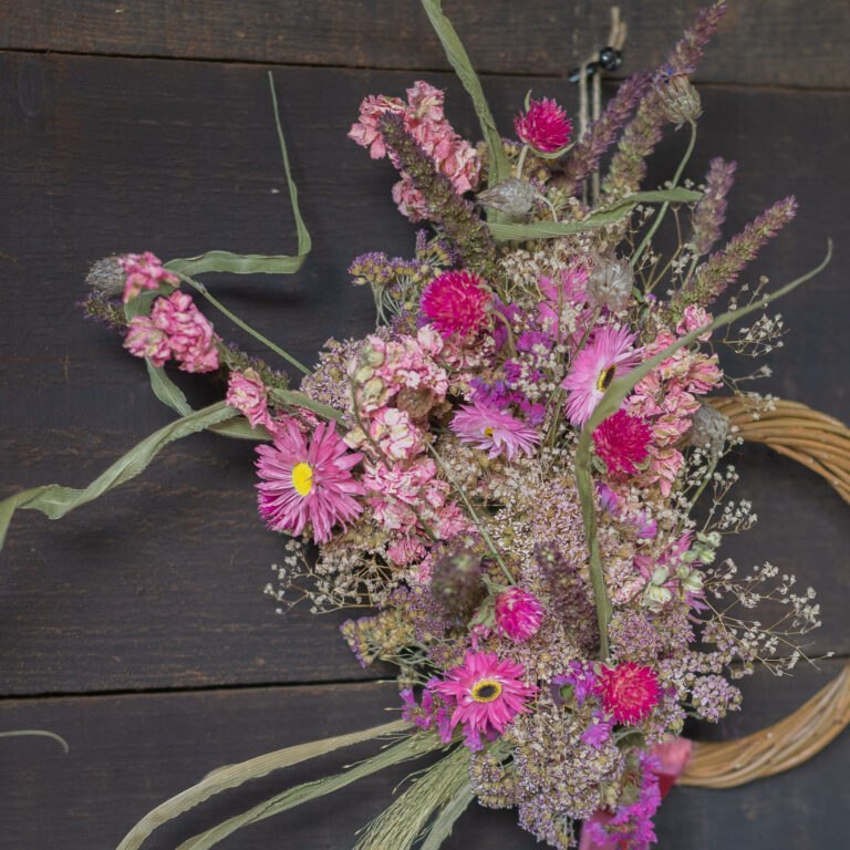 A Mini Wreath - Green and White featuring pink and purple flowers with greenery is displayed on a dark wooden wall.