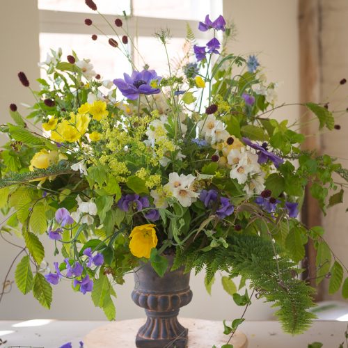The Flower Club arrangement in a dark vase features vibrant yellow, white, and purple blooms with lush green foliage on a light-coloured table.
