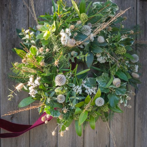 A Christmas wreath crafted in our course features green leaves, small white and brown flowers, and sprigs of grass. It adorns a wooden door with a red ribbon affixed at the bottom.