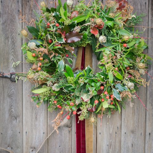 A verdant wreath adorned with green leaves, red berries, and natural accents graces a rustic wooden door, showcasing the skills taught in our Christmas Wreath-Making Course.
