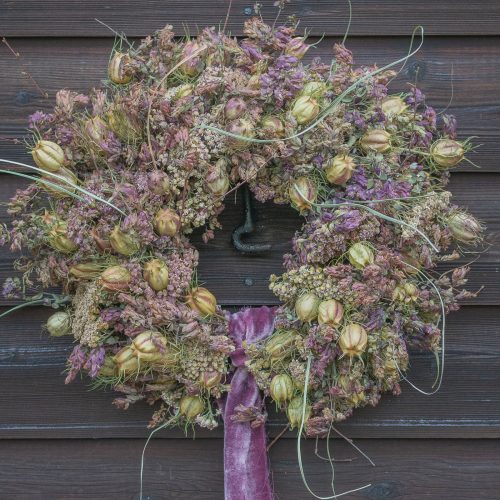 The "Half Moon" Everlasting Wreath, featuring dried purple and green flowers, is elegantly displayed on a wooden wall with a purple ribbon detail.