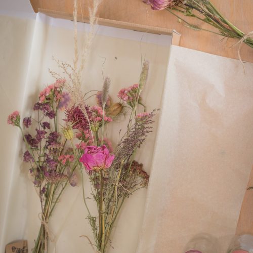 A bouquet of Letterbox Flowers in pink, purple, and beige hues is displayed on paper with two glass jars beside it on a wooden surface.