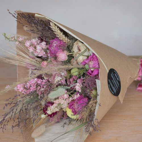 The "Half Moon" Everlasting Wreath, featuring pink roses, purple accents, and various grasses, is wrapped in brown paper and placed on a wooden surface.