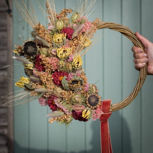 A hand showcases the "Half Moon" Everlasting Wreath, a circular woven design featuring dried flowers and grasses in red, yellow, and brown, adorned with a red ribbon against a pale green wooden wall.