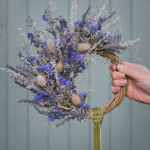 A hand showcases the "Half Moon" Everlasting Wreath, crafted from dried purple and white flowers with a green ribbon, set against a light grey background.