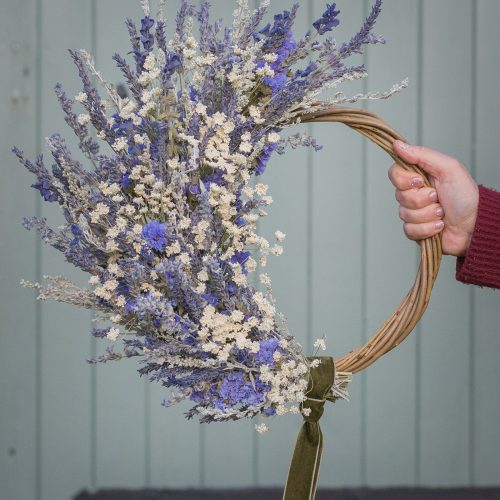 A hand presents the "Half Moon" Everlasting Wreath, showcasing a circular arrangement of dried lavender and white flowers on a rustic wooden hoop, set against a light green backdrop.