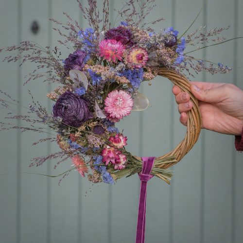 A hand holds the "Half Moon" Everlasting Wreath, adorned with dried flowers and grasses, featuring a purple ribbon cascading down.