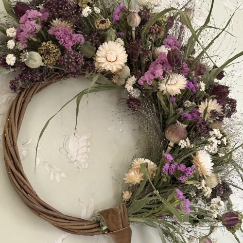 The "Half Moon" Everlasting Wreath, with purple, white and cream dried flowers, hangs on the wall from a brown velvet ribbon.