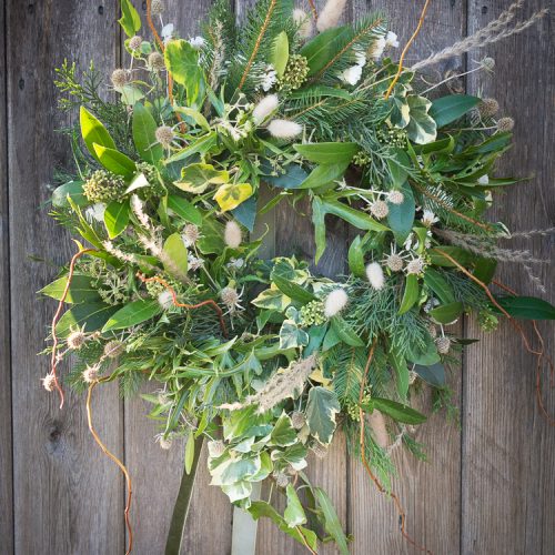 A Christmas wreath, lush with various green leaves and adorned with small white flowers, hangs on a wooden wall.