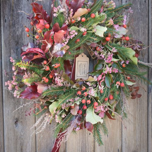 A Christmas wreath featuring red leaves, pine branches, pink flowers, berries, and a small tag hangs on a rustic wooden door.