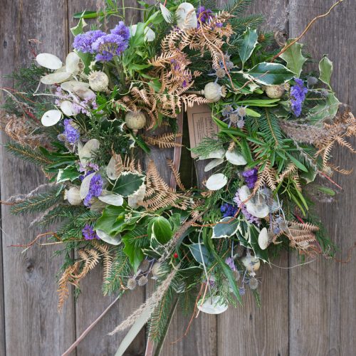 The Christmas Wreath showcases a rustic blend of mixed greenery, dried flowers, and leaves set against a wooden background.