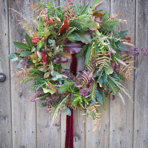 A Christmas wreath with rustic charm features green leaves, ferns, and purple flowers on a wooden door.