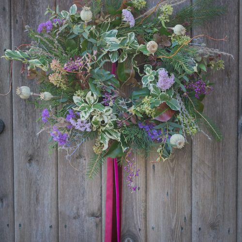 A Christmas wreath with greenery and purple flowers, accented by a long pink ribbon, adorns a wooden door.