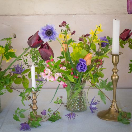 A table adorned with brass candle holders and white candles displays vibrant purple, red, and yellow blooms from the "Design Workshop: Creating Wedding Flowers" against a light background.
