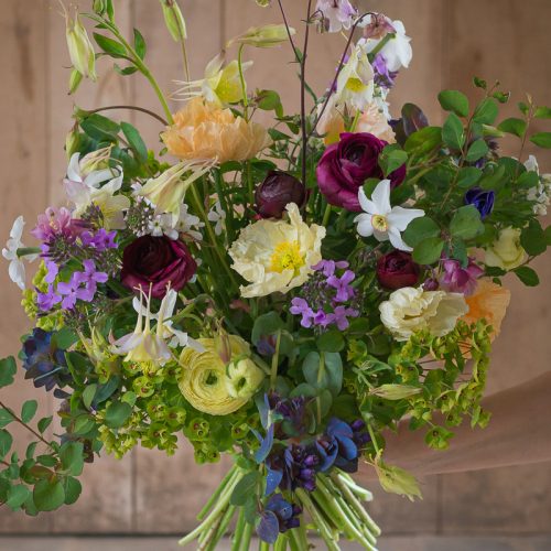 A hand presents a "Design Workshop: From Field To Vase" bouquet with roses, peonies, and greenery against a wooden backdrop.