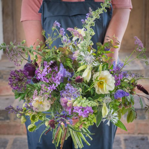 A person wearing overalls and a pink shirt holds a large bouquet from "Design Workshop: From Field To Vase," featuring purple, white, and yellow flowers, set against a blurred brick backdrop.