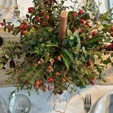 A table laid with plates and cutlery displays the Festive Table Centrepiece Course: greenery, red berries, dried flowers, and a tall brown candle on a pale tablecloth.