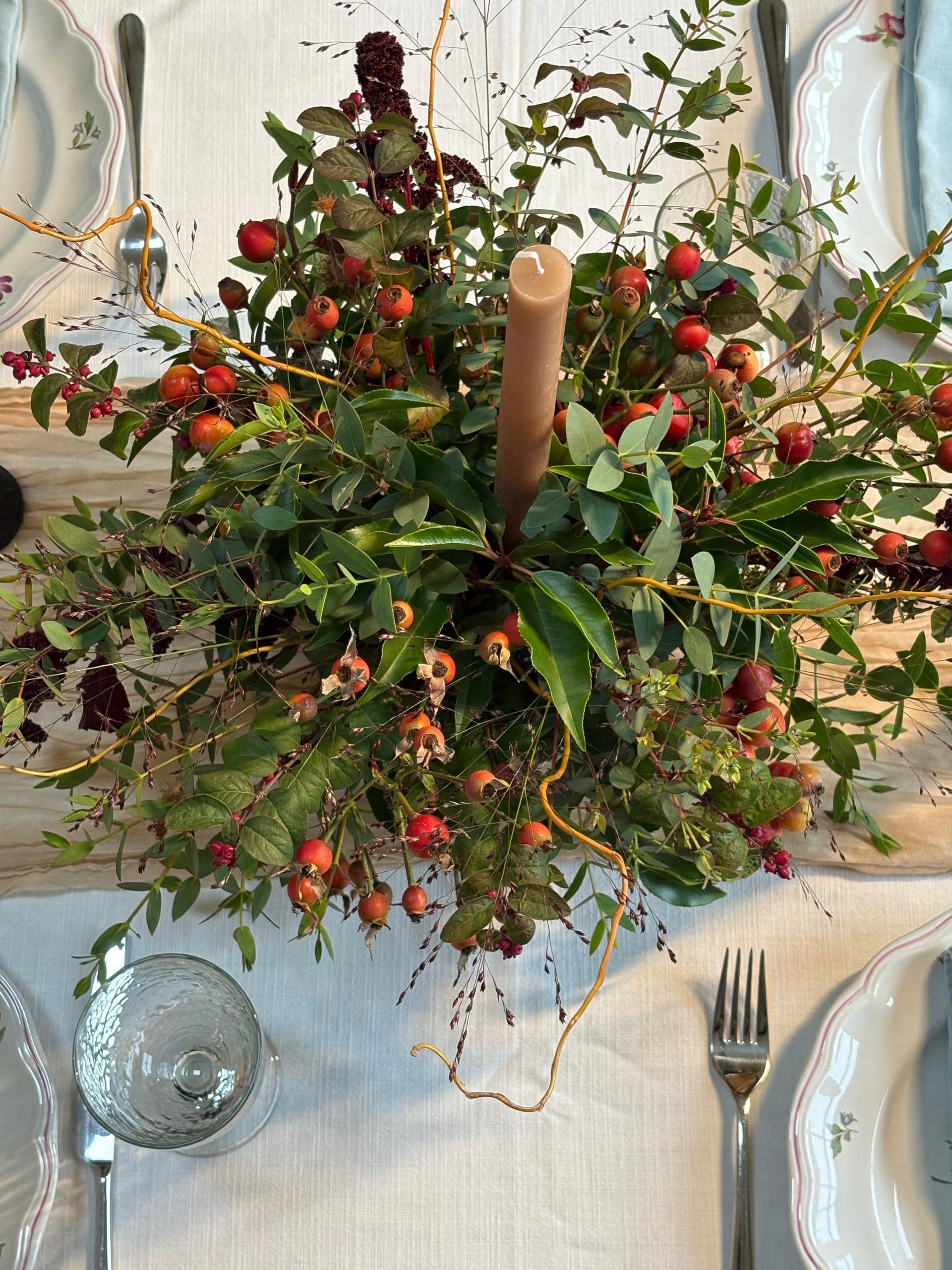 A table laid with plates and cutlery displays the Festive Table Centrepiece Course: greenery, red berries, dried flowers, and a tall brown candle on a pale tablecloth.