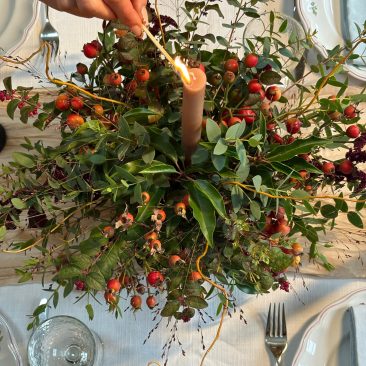 A hand lights a brown candle in the Festive Table Centrepiece Course, with green leaves and red berries on a table set with crockery, cutlery, and glasses.