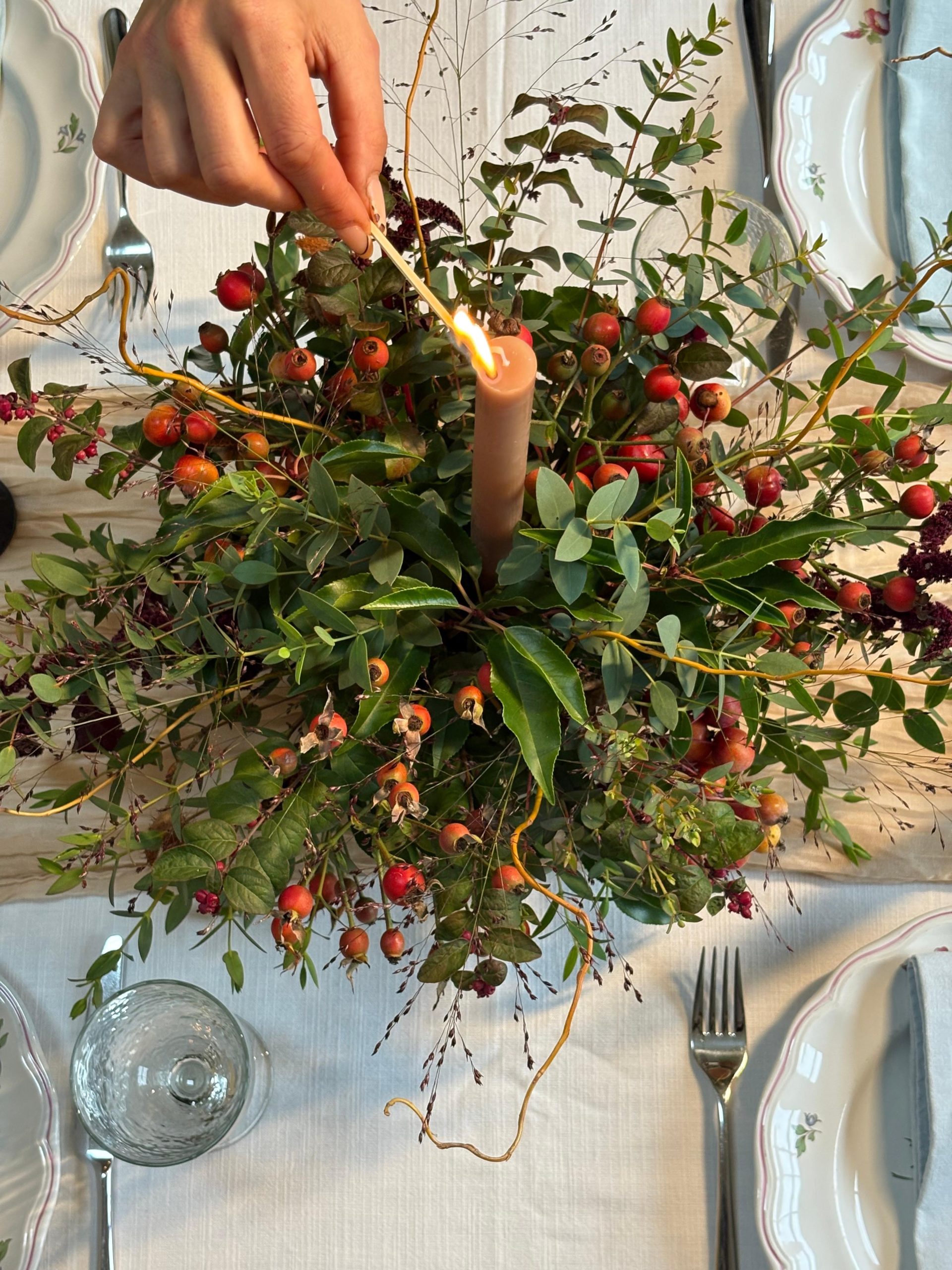 A hand lights a brown candle in the Festive Table Centrepiece Course, with green leaves and red berries on a table set with crockery, cutlery, and glasses.